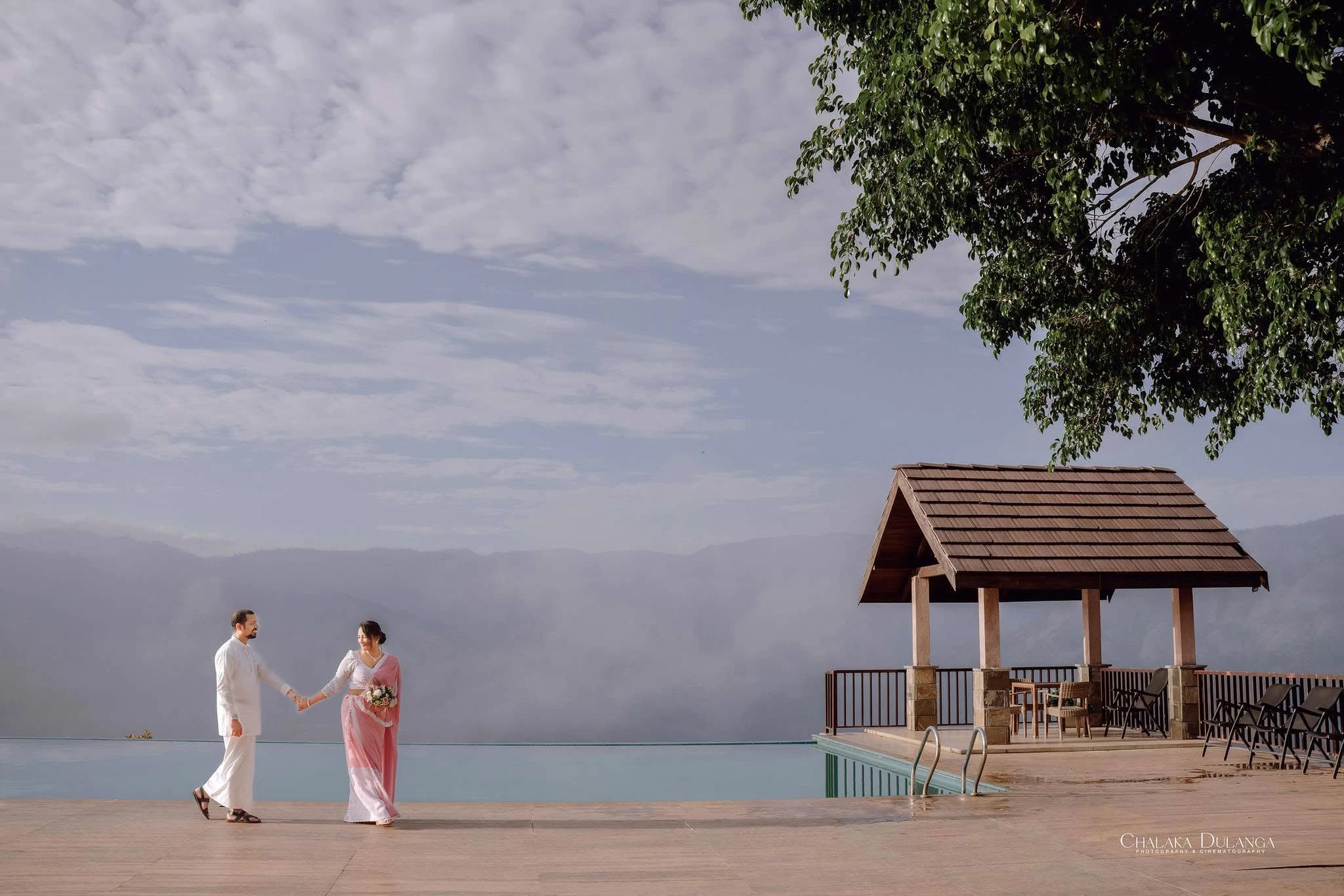 A couple walking hand in hand beside an infinity pool with a pavilion and misty mountains